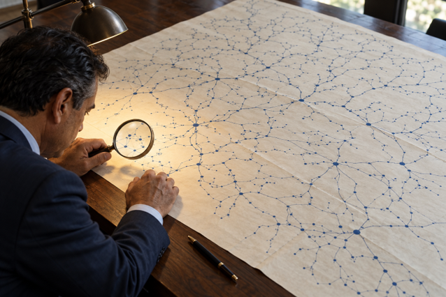 Empresario observa con una lupa una pequeña zona de un mapa amplio de conexiones sobre una mesa.
